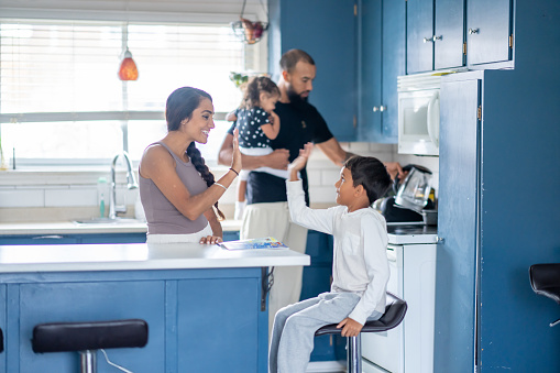 A young Mother stands at her kitchen island as she helps her son with his homework.  The two are dressed comfortably and giving each other a high five as the Father holds his daughter in his arms and works away in the background.