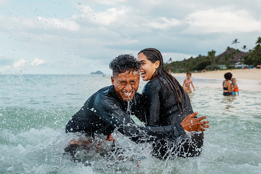 A young man of Pacific Islander ethnicity smiles and he plays with his female partner, splashing each other in the ocean during a tropical vacation.