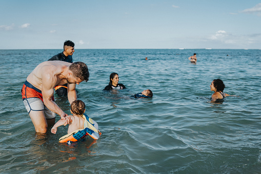 A loving Caucasian father stands in the shallow ocean and plays with his Eurasian one year old son while at the beach with family and friends in Hawaii. The father is helping his son take off his life jacket.