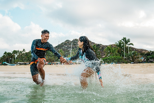 Pacific Islander man and Hispanic wife playfully splash in the ocean while at the beach in Hawaii.