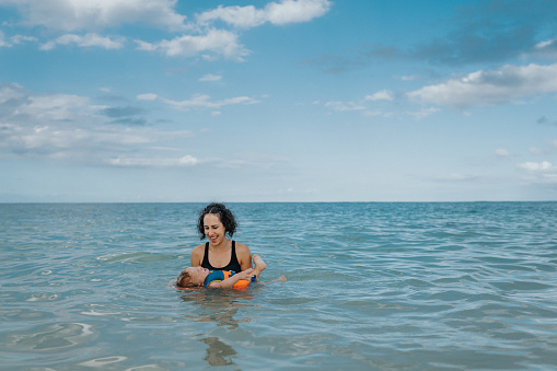 An Eurasian one year old boy, wearing a lifejacket, lays on his back in the ocean while his mother of Pacific Islander holds him up. The family is on vacation in Hawaii.
