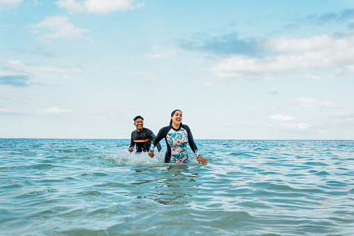 A pretty Hispanic young woman laughs as her husband of Pacific Islander descent playfully chases her through shallow water at the beach in Hawaii.