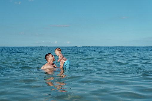 A Caucasian man affectionately holds and looks lovingly at his one year old Eurasian son as they swim together in the ocean while on vacation in Hawaii.