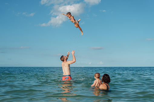 A Caucasian man playfully throws his Eurasian daughter up into the air while swimming in the ocean. The family is enjoying time at the beach in Hawaii with family and friends.