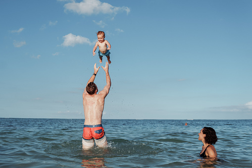 A Caucasian man who is at the beach in Hawaii with his family, playfully throws his Eurasian toddler son up into the air while his wife swims nearby and watches.