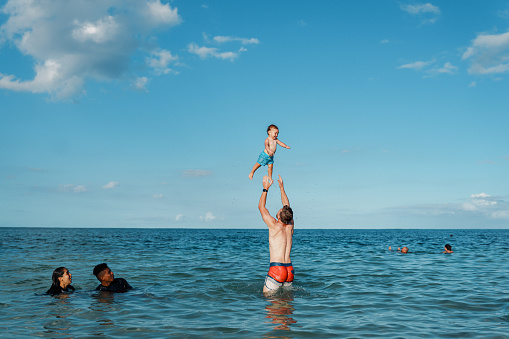 A Caucasian man playfully throws his Eurasian toddler son up into the air while swimming in the ocean. The family is enjoying time at the beach in Hawaii with family and friends.
