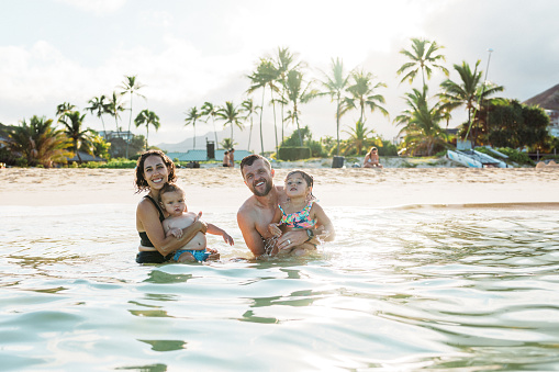A beautiful multiracial woman of Pacific Islander descent and her Caucasian husband smile directly at the camera and affectionately hold their one year old son and three year old daughter while swimming at the beach in Hawaii.