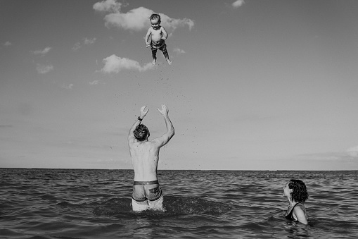 Black and white photo of a Caucasian man who is at the beach in Hawaii with his family, playfully throwing his Eurasian toddler son up into the air while his wife swims nearby and watches.