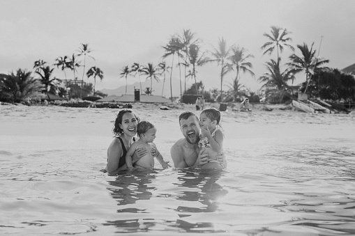 A beautiful multiracial woman of Pacific Islander descent and her Caucasian husband smile directly at the camera and affectionately hold their one year old son and three year old daughter while swimming at the beach in Hawaii.
