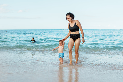 A beautiful multiracial woman of Pacific Islander descent holds hands with her one year old Eurasian son and walks along the beach while on vacation in Hawaii.