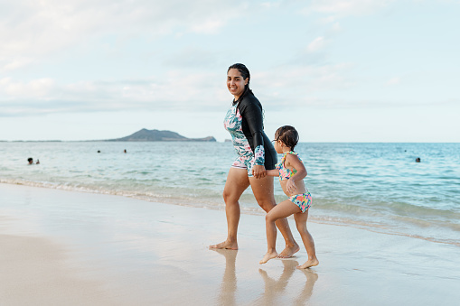 A young woman of Hispanic descent smiles and holds hands with her three year old niece as they walk in the sand after swimming in the ocean together in Hawaii.