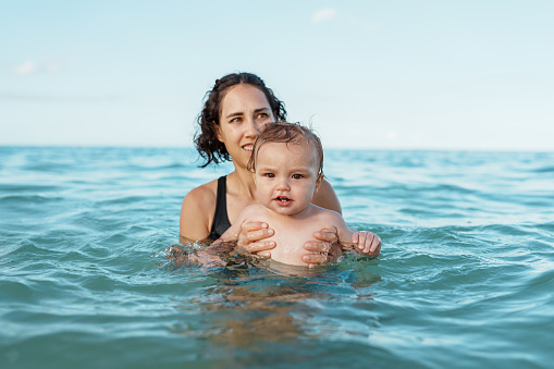 A beautiful multiracial woman of Pacific Islander descent holds her one year old Eurasian son while swimming in the ocean during a family vacation in Hawaii.