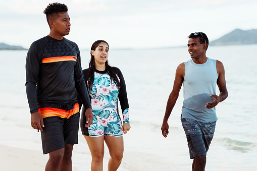 An Hispanic woman holds hands with her husband of Pacific Islander descent as they walk along the beach and cheerfully talk with a friend of Indian descent. The group of friends live in Hawaii and are enjoying quality time together outdoors while also being active.