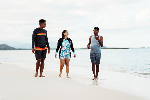 An Hispanic woman holds hands with her husband of Pacific Islander descent as they walk along the beach and cheerfully talk with a friend of Indian descent. The group of friends live in Hawaii and are enjoying quality time together outdoors while also being active.