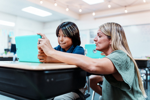 A young boy of Asian ethnicity sits at a classroom desk as his female teacher squats next to him, describing how to use a tablet for schoolwork.