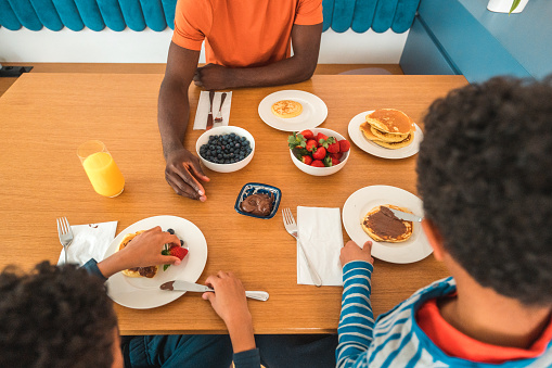 Un unrecognizable father and his two young sons gather around the table for a delightful breakfast. They are eating pancakes and fresh fruit.