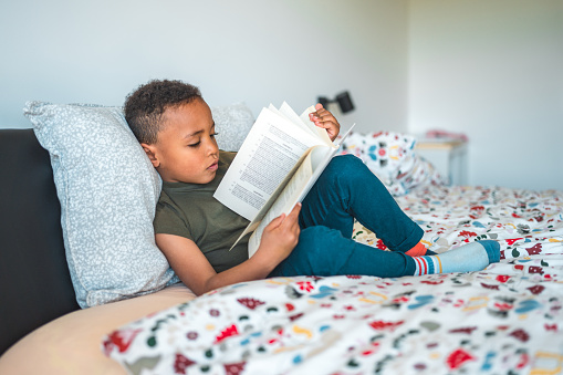 Adorable Mixed race preschooler boy sitting on a bed in a domestic bed room reading a book.