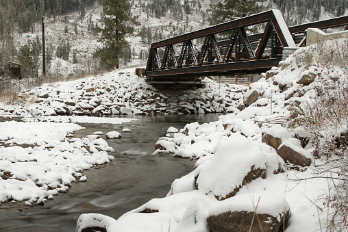 An old, snow covered, iron bridge over a slow moving creek, with ice and snow covering the ground.