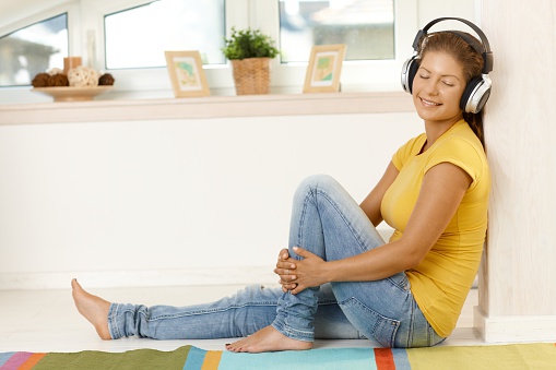 Happy young woman sitting on floor at home, listening to music on headphones, smiling, eyes closed...