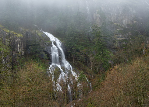 Long exposure of a mountain waterfall coming down across a sheer rock face, surrounded by fog rolling in and low lying trees.