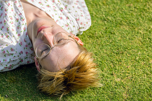 Elevated view of young woman laying on the grass with her eyes closed on sunny day, background with copy space, full frame horizontal composition