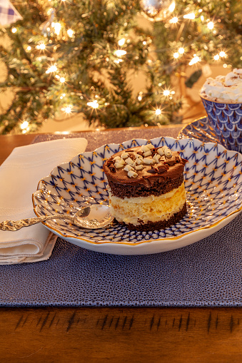 Hot chocolate and layered dark and white chocolate dessert on a fancy bone China plate tipped with gold and a silver spoon in front of a Christmas tree.