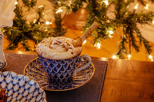 Cup of Hot chocolate in a fancy bone China cup with gold and a silver spoon in front of a Christmas tree.