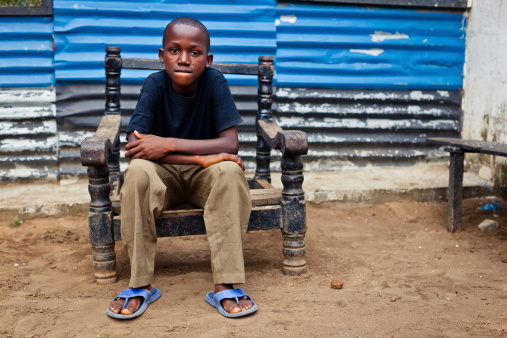 An African boy sitting in a broken chair with his hands on his knees while looking seriously at the camera.