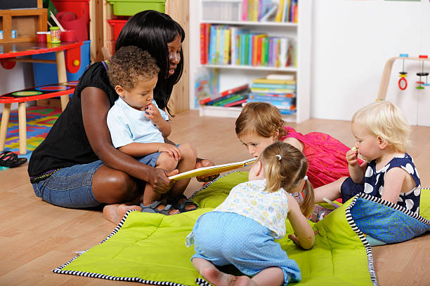 childminder/accompagnatore lettura a un gruppo di bambini in camera dei bambini - baby1 foto e immagini stock