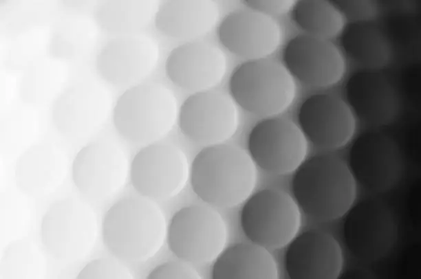 A macro shot of a plastic golf ball. The ball is white and the surface is covered in round white indentations. There’s a strong shadow across the ball from left to right where a strong side falls across it. The round white dimples on the surface are identical in shape and size.