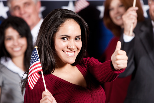 Very happy 20-25  Hispanic female waving small American flag and making thumbs up gesture to viewer.  Group of pleased supporters look on from behind.