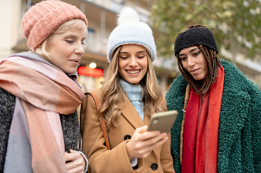 Three women bundled up in winter attire, sharing and enjoying content on a smartphone while standing on a city sidewalk, reflecting connectivity and friendship in the digital age.