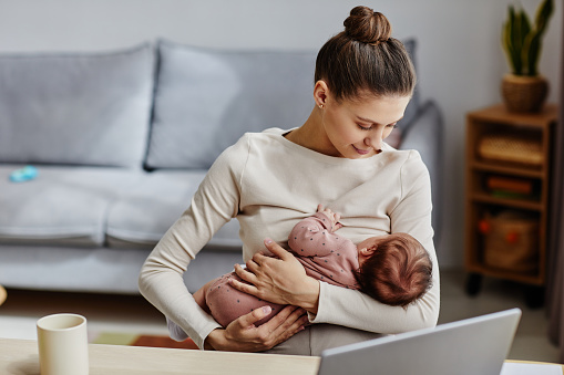 Waist up shot of happy Caucasian mother sitting by computer and lulling her baby during work break at home, copy space