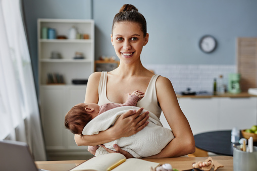 Waist up portrait of beaming Caucasian mother looking at camera and sitting at table with baby in arms, copy space