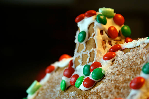 Sweet gingerbread house decorated with icing details and colorful candies, with blurred Christmas lights behind it. Warm festive mood perfect for holiday backgrounds and seasonal design.