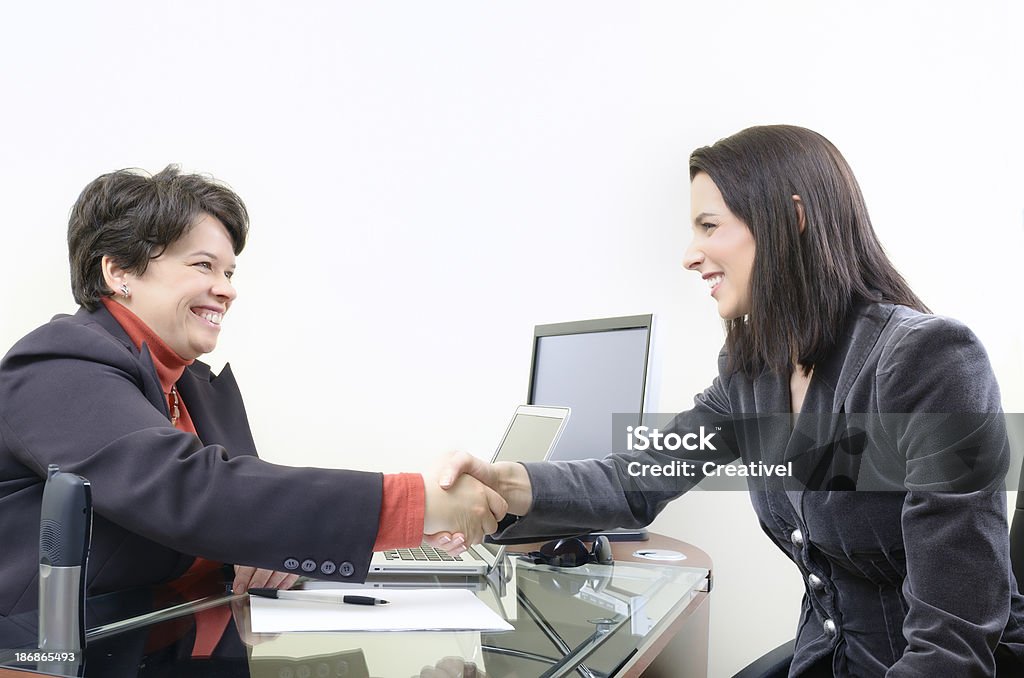 Two businesswomen shaking hands "Businesswomen shaking hands across desk in office, smiling" 30-39 Years Stock Photo