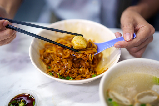 The cropped shot captures an Asian woman savoring flat rice fish ball noodles soup with dark soy sauce at a Penang hawker stall.