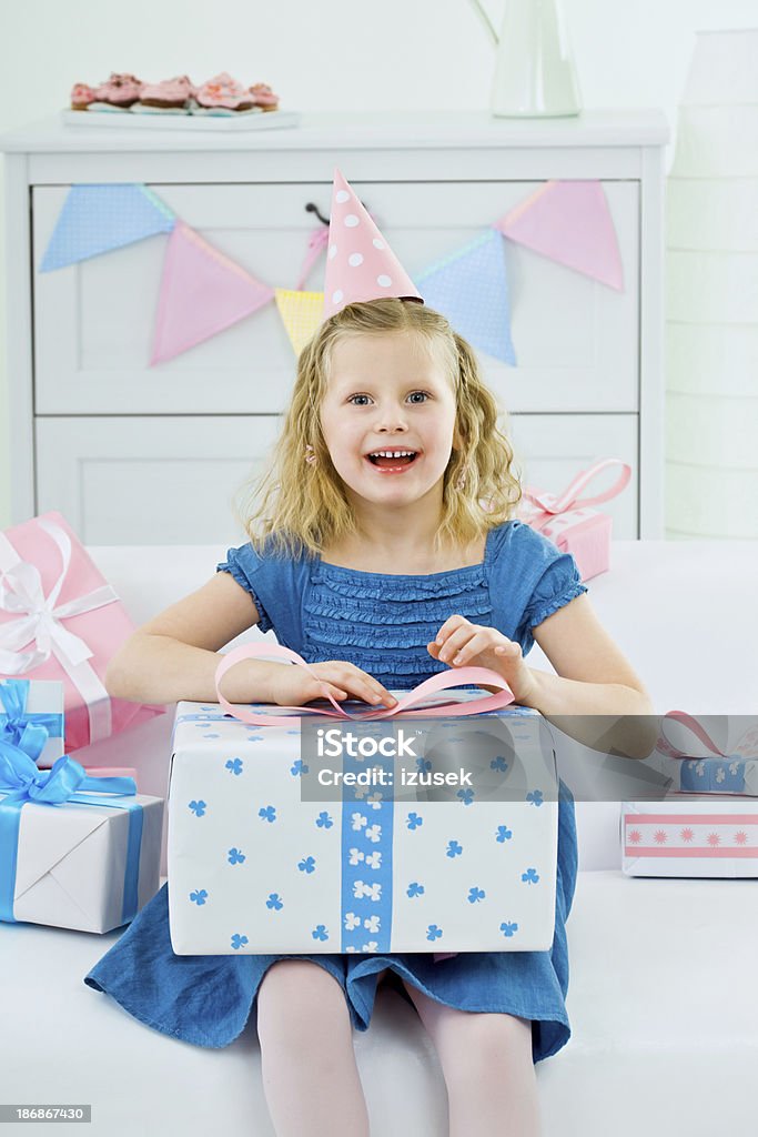 Cute girl with birthday present "Cute little girl wearing party hat sitting on sofa among birthday presents and holding the big one on her legs, looking at camera and laughing" 6-7 Years Stock Photo