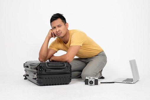 A sad and bored Asian man is sitting on an isolated white studio background with his belongings and luggage. missed the flight, boredom, no holiday