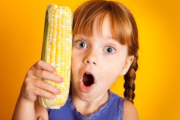 Color photo of a shocked little red-haired girl holding corn on the cob.