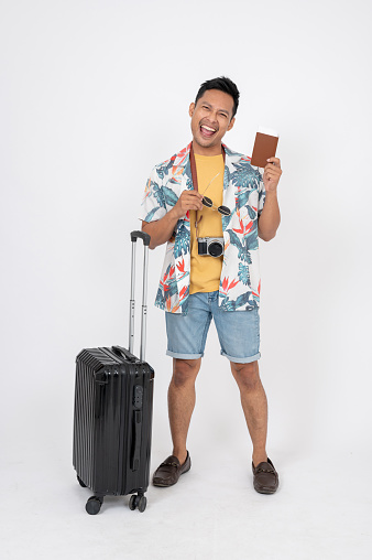 A cheerful Asian man in summer clothes is standing on an isolated white studio background with his luggage, showing his passport, excited for his summer vacation.