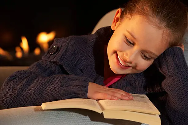 Portrait 7 year old girl reading by firelight on sofa