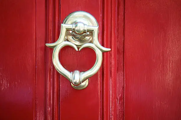 Golden brass door knocker in the shape of a heart shines on bright enamel red door