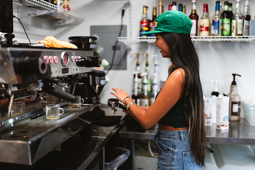 A young adult female barista of Asian descent stands in the kitchen of a food truck, preparing coffee drinks.