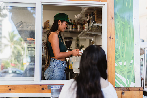 Rear view of a young woman standing outside, ordering a beverage from a woman of Asian ethnicity who is working in a coffee truck.