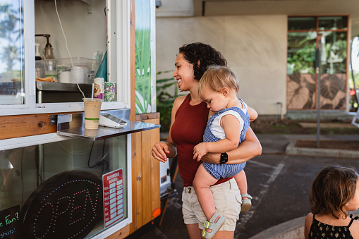 A young woman of Eurasian ethnicity holds her multiracial toddler son while standing outside, ordering coffee from an outdoor food truck.
