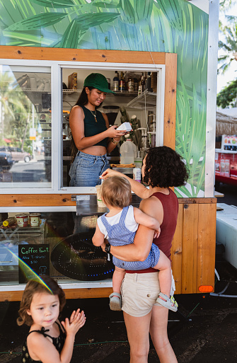 A young woman of Eurasian ethnicity holds her multiracial toddler son while standing outside, paying a barista at a food truck while toddler daughter stands beside her.