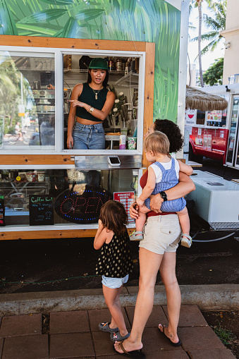 A young female barista of Asian ethnicity stands in a food truck, speaking to a female customer and her toddler children outside on a beautiful day.