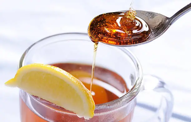 A spoonful of honey drips into a cup of hot tea with lemon.