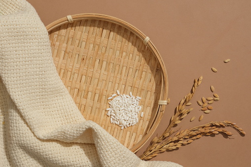 Flat winnowing basket, white rice, whole grain rice and beige fabric displayed on a brown background. Ideal space for advertising new products.